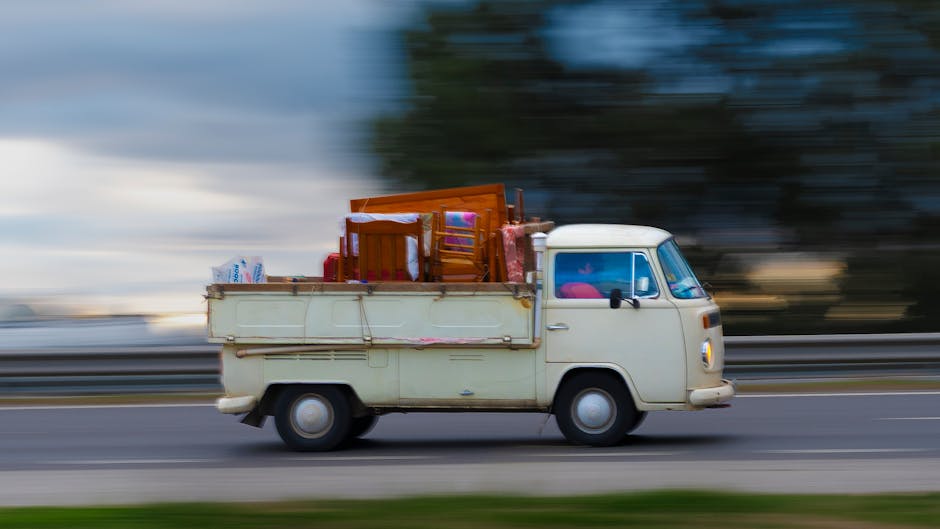 A vintage cream-colored van is shown in motion on a highway, with a blurred background indicating speed. The open cargo area at the back of the van is loaded with various household items, including wo