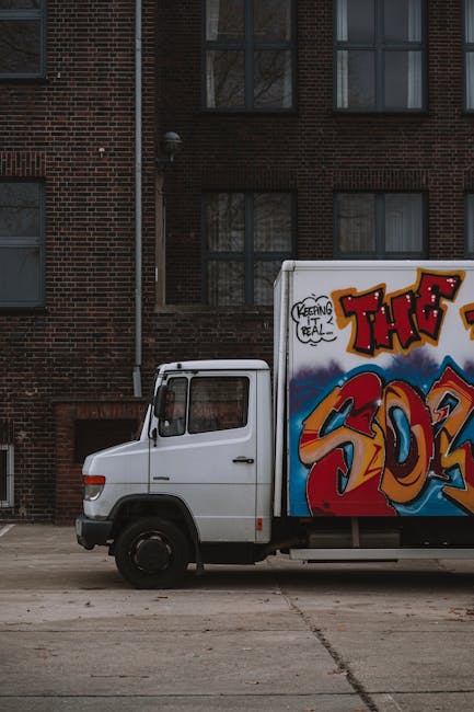 A white moving van parked on a city street during daylight hours, with a driver sitting in the cab, visible through the open window. The van bears the text 'MOVING COMPANY LOCAL & LONG DISTANCE' on it