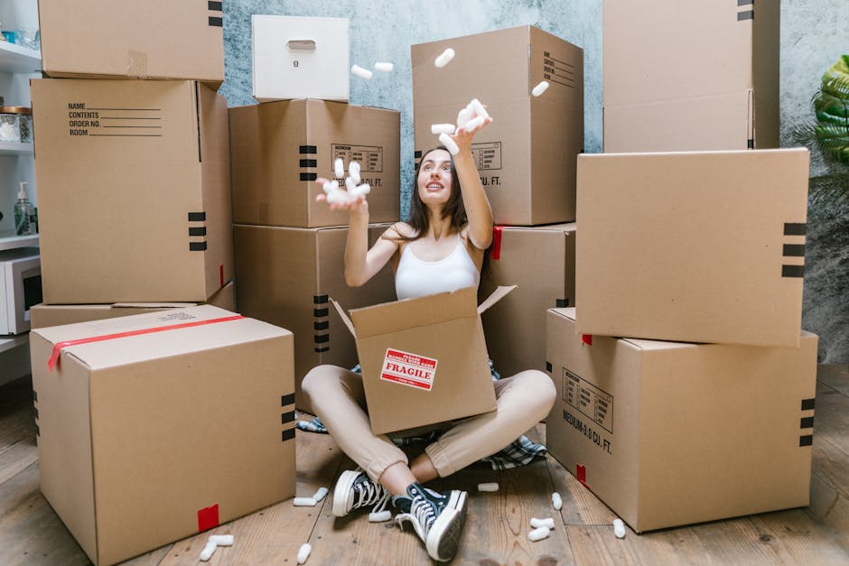 A young man with a headband and casual clothing, sitting on a wooden floor inside a room with large, arched windows, surrounded by several cardboard boxes sealed with red tape, indicating a home reloc
