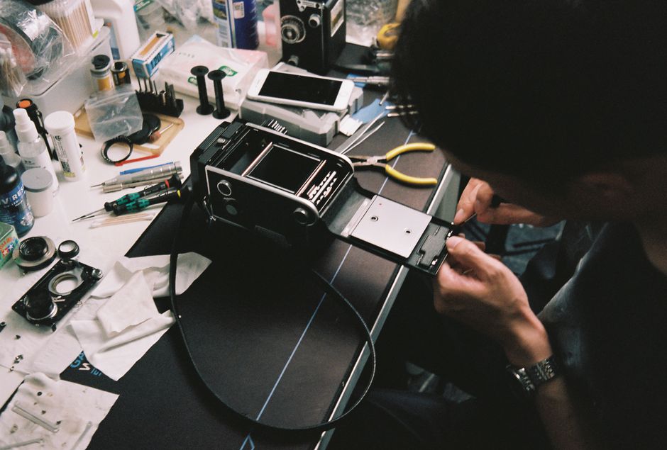 A person wearing glasses and a wristwatch is working on a disassembled electronic device, possibly a tablet or laptop, placed on a black work surface within a cluttered workspace. The workspace contai