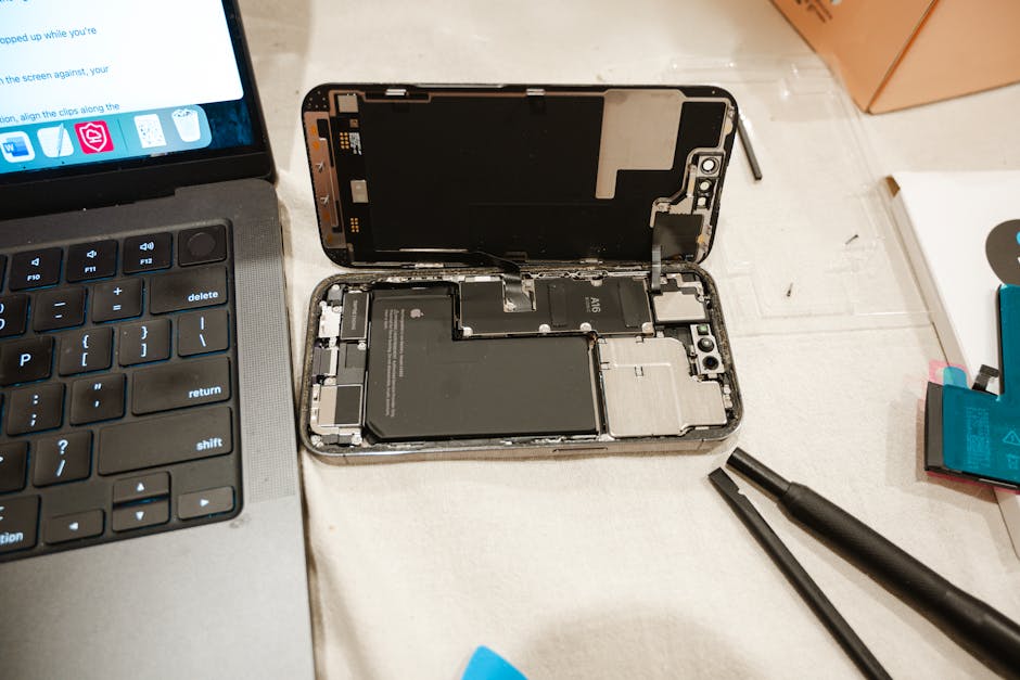 A person wearing glasses and a wristwatch is working on a disassembled electronic device, possibly a tablet or laptop, placed on a black work surface within a cluttered workspace. The workspace contai
